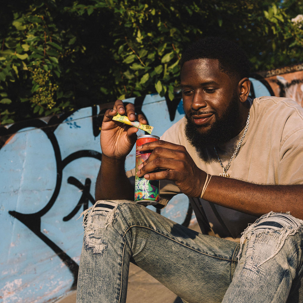 A man sitting outdoors on a bench holding a can of hemp THC-infused soda and a single Lemon Drop mix stick.
