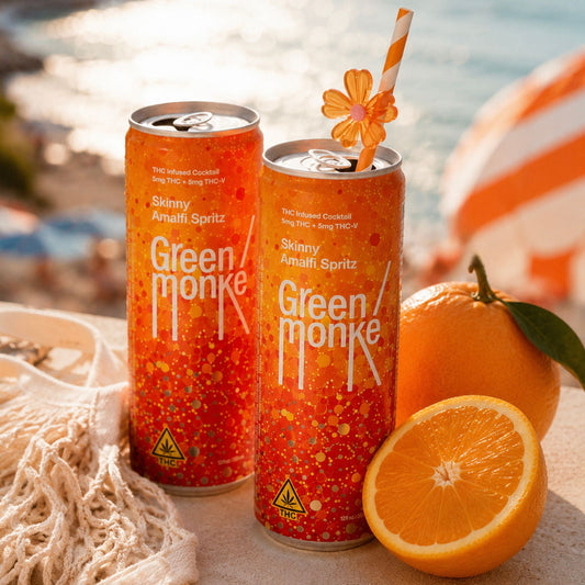 Two cans of Green Monke Skinny Amalfi Spritz THC & THCV cocktails are displayed on a beach table with oranges, a net, and a striped umbrella in the background.